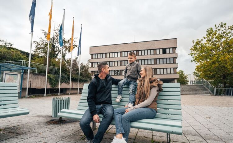 Eine dreiköpfige Familie sitzt auf einer Bank vor dem Rathaus und unterhält sich: Klick öffnet eine vergrößerte Ansicht