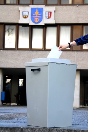 Symbolbild Wahl, das eine Wahlurne vor dem Rathaus zeigt in die ein Stimmzettel eingeworfen wird