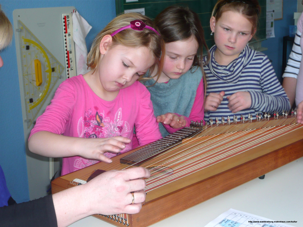 Drei Mädchen sehen sich eine Zither der Musikschule an Drei Mädchen sehen sich eine Zither der Musikschule an
