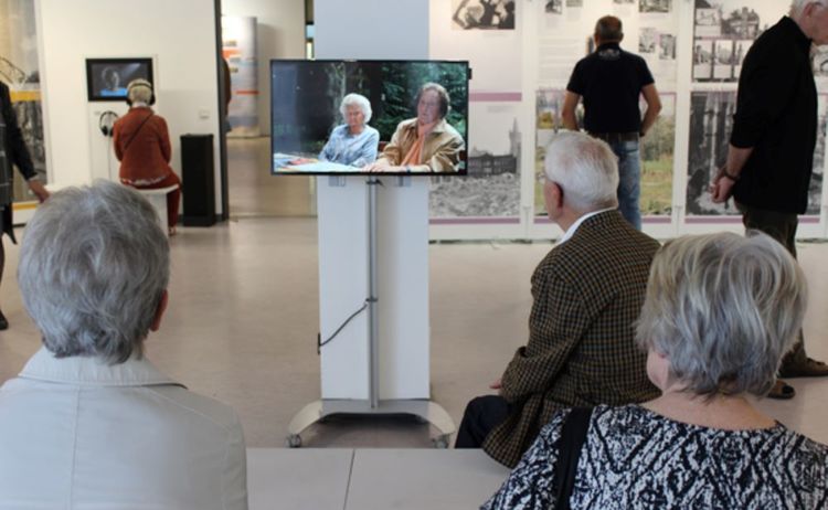 Besucher sehen sich auf einem Fernseher ein Interview der Ausstellung Verschwundene Orte an: Klick öffnet eine vergrößerte Ansicht