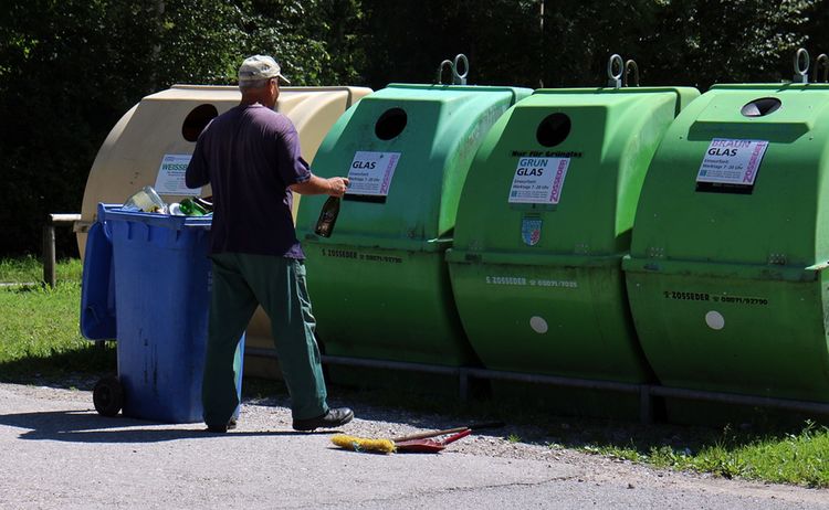 ein Mann trägt eine leere Flasche zu einem Glascontainer. Es stehen insgesamt 4 Glascontainer und eine blaue Tonne in der Wiese: Zum Vergrößern auf Bild klicken